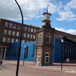Historisch gebouw met klok en moderne architectuur in een stadscentrum, naast een kanaal en wandelpad.