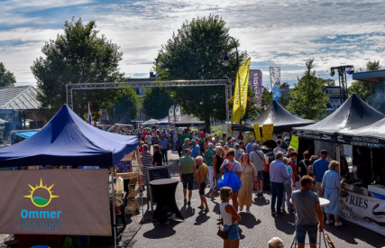 Drukke markt in de buitenlucht met kraampjes en mensen onder een blauwe lucht tijdens een zonnige dag.