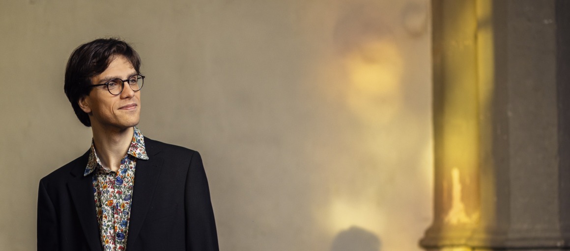 Man in zwart pak met kleurrijk hemd en bril kijkt opzij, staand naast een muur met lichteffecten.