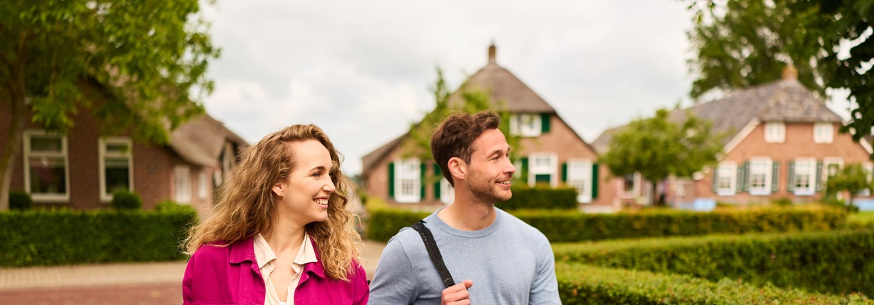 Twee jonge volwassenen lopen vrolijk buiten in een rustig dorp, de vrouw trekt een koffer achter zich aan.