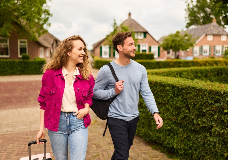 Twee jonge volwassenen lopen vrolijk buiten in een rustig dorp, de vrouw trekt een koffer achter zich aan.