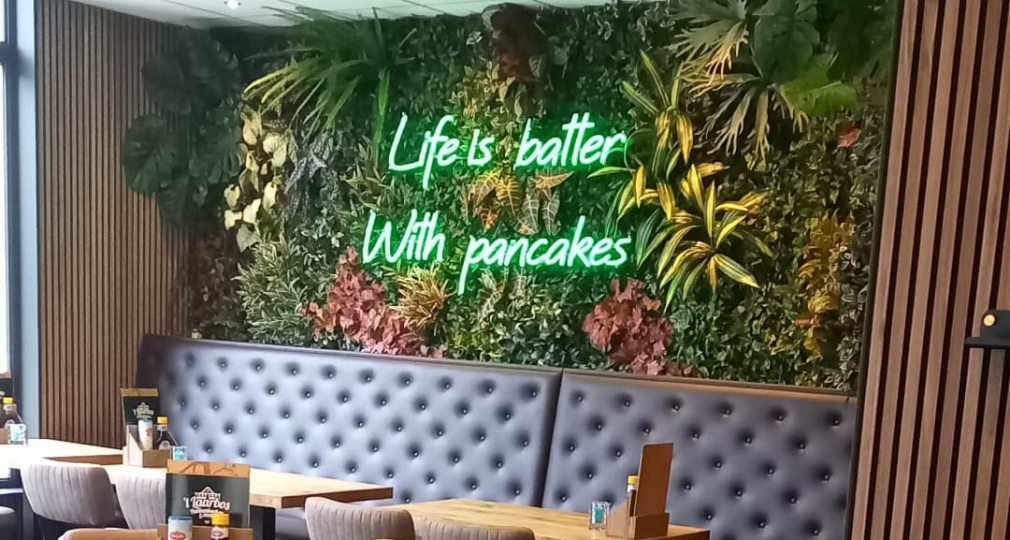 Gemütliches Café mit Holztischen, gepolsterten Stühlen, Pflanzenwand und leuchtendem Neon-Schriftzug.
