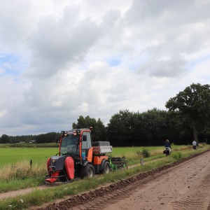 Een tractor staat geparkeerd langs een landweg met groene velden en enkele mensen op de achtergrond