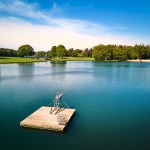 Een rustig meer met een houten vlot en duikplank, omringd door groene bomen en een heldere lucht.