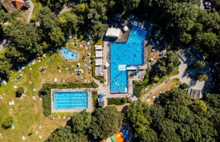 Luchtfoto van een druk buitenzwembadcomplex, omgeven door grasvelden, bomen en mensen die zonnebaden.