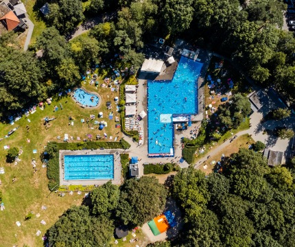 Luchtfoto van een druk buitenzwembadcomplex, omgeven door grasvelden, bomen en mensen die zonnebaden.