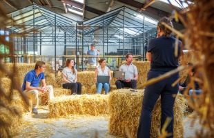 Een groep mensen zit op hooibalen tijdens een informele vergadering in een kas, met laptops en notities.
