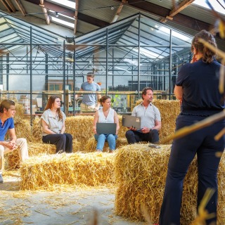 Een groep mensen zit op hooibalen tijdens een informele vergadering in een kas, met laptops en notities.