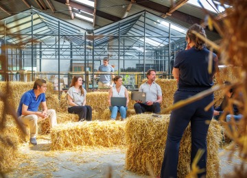 Een groep mensen zit op hooibalen tijdens een informele vergadering in een kas, met laptops en notities.
