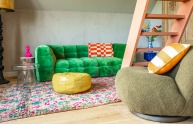 Eigenzinnig interieur met groene bank, kleurrijk vloerkleed, poef en opvallende trap in een woonkamer.