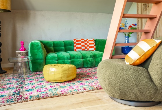 Eigenzinnig interieur met groene bank, kleurrijk vloerkleed, poef en opvallende trap in een woonkamer.