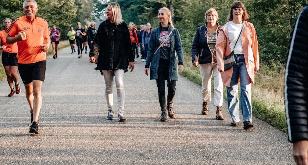 Een groep mensen wandelt en jogt op een rustige weg omringd door bomen tijdens een zonnige dag.