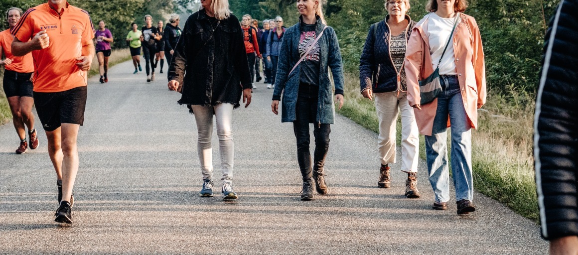Een groep mensen wandelt en jogt op een rustige weg omringd door bomen tijdens een zonnige dag.