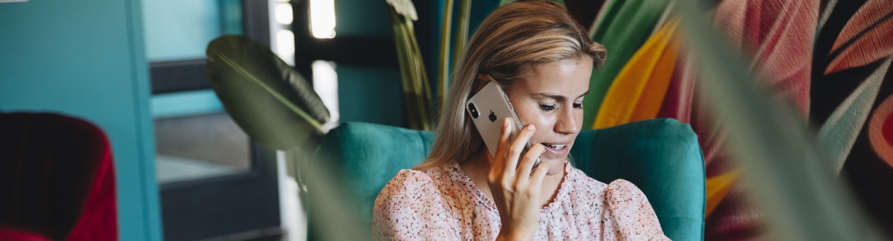 Vrouw zit in een turquoise stoel, telefoneert met een smartphone en werkt op een laptop in een kleurrijke ruimte.