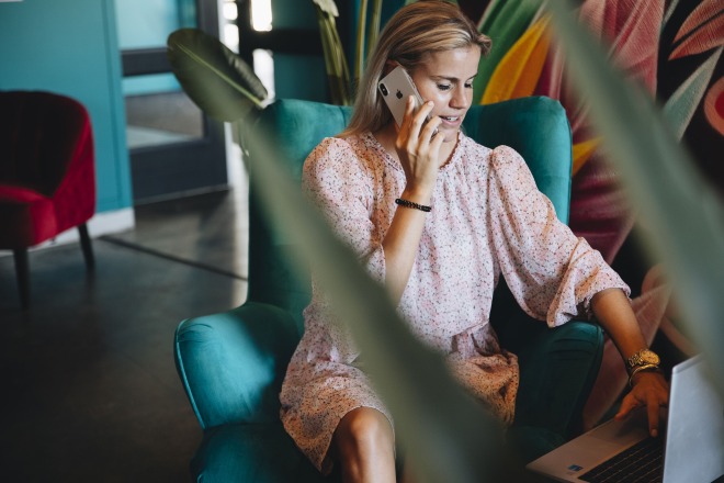 Vrouw zit in een turquoise stoel, telefoneert met een smartphone en werkt op een laptop in een kleurrijke ruimte.