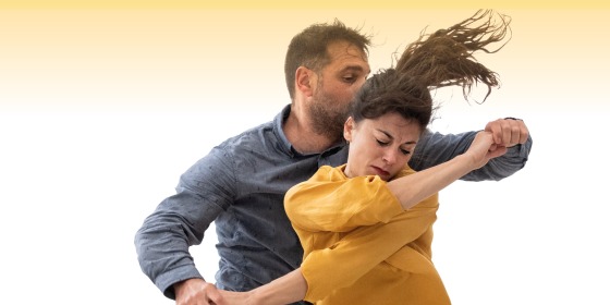 Twee mensen dansen expressief samen, waarbij de vrouw een gele blouse draagt en de man een blauwe overhemd.