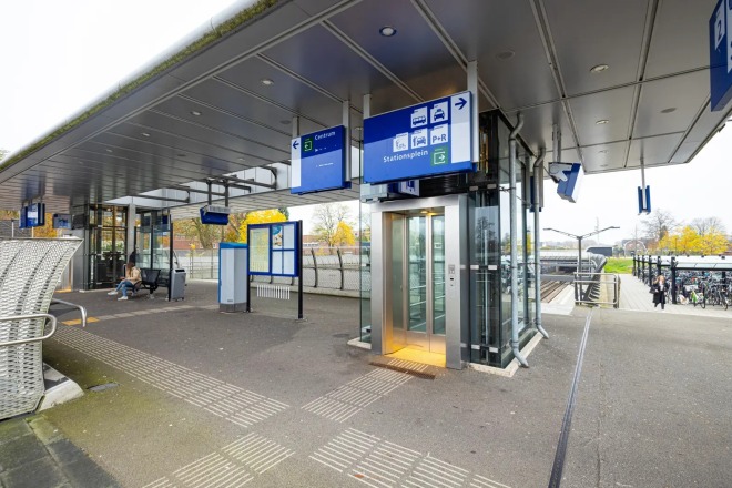 Modern stationsplein met overdekte wachtruimte, lift, fietsenstalling en duidelijke bewegwijzering.