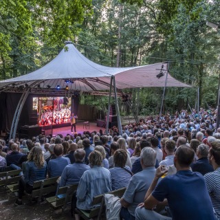 Openluchttheater in het bos met publiek dat naar een optreden kijkt onder een grote tentconstructie.