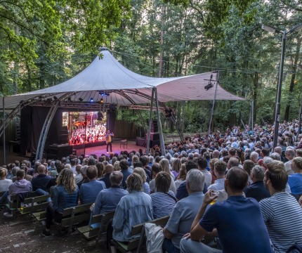 Openluchttheater in het bos met publiek dat naar een optreden kijkt onder een grote tentconstructie.