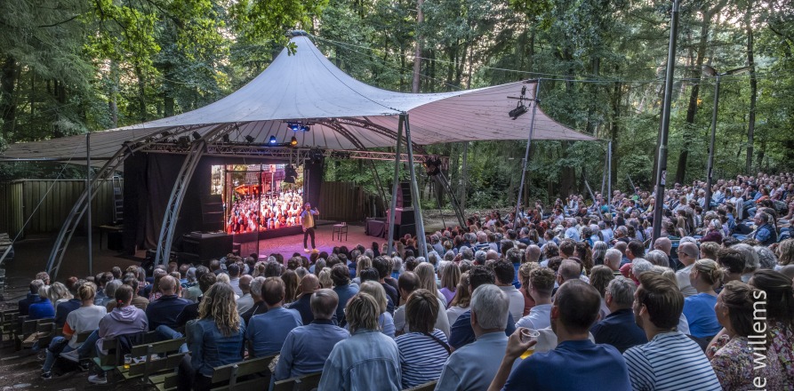 Outdoor theater in a forest with an audience watching a performer on stage under a large canopy tent.