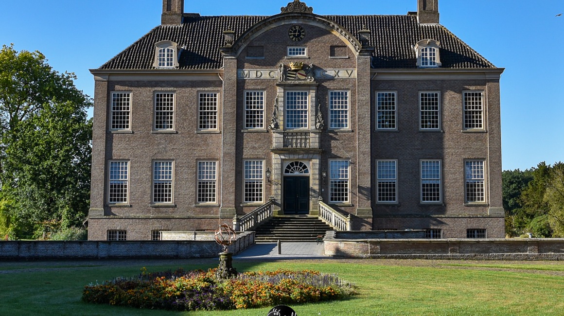 Vooraanzicht van een historisch Nederlands landhuis met grote ramen en tuin op een zonnige dag.