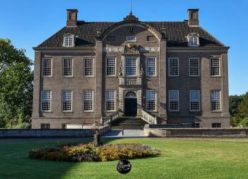 Vooraanzicht van een historisch Nederlands landhuis met grote ramen en tuin op een zonnige dag.