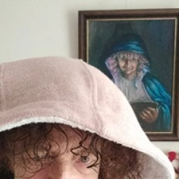 Vrouw met krullend haar en roze capuchonposeert voor een schilderij waarop een lachende vrouw met blauwe hoed staat.