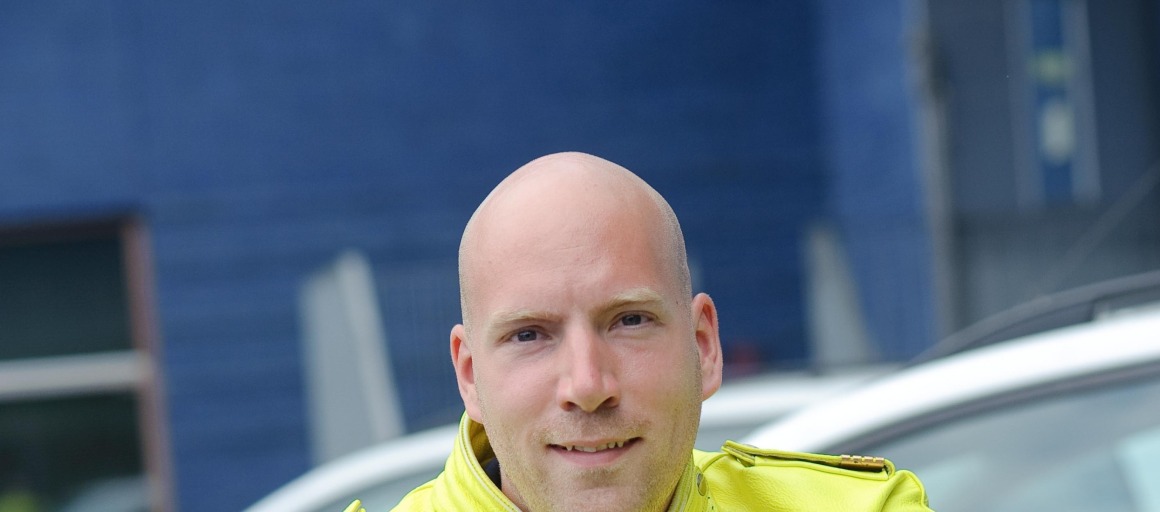 Politieagent in gele jas leunend op motorfiets, met politievoertuigen op de achtergrond.