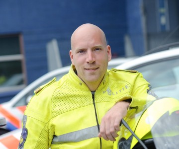 Politieagent in gele jas leunend op motorfiets, met politievoertuigen op de achtergrond.