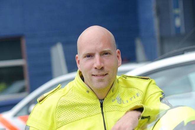 Politieagent in gele jas leunend op motorfiets, met politievoertuigen op de achtergrond.