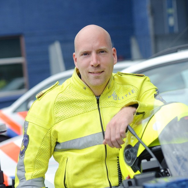 Politieagent in gele jas leunend op motorfiets, met politievoertuigen op de achtergrond.