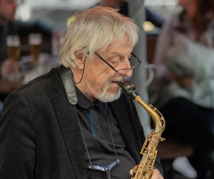 Oudere man met grijs haar speelt saxofoon in een café, bril op de neus, mensen op de achtergrond.