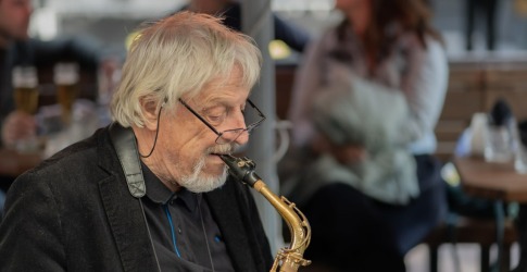 Oudere man met grijs haar speelt saxofoon in een café, bril op de neus, mensen op de achtergrond.