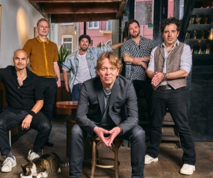 Een groep van zes mannen poseert samen binnen; een kat eet uit een bakje op de houten vloer.