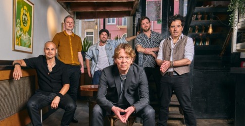 Een groep van zes mannen poseert samen binnen; een kat eet uit een bakje op de houten vloer.