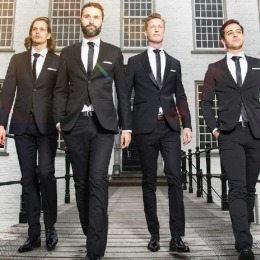 Vier mannen in pakken lopen over een brug voor een wit gebouw met grote ramen, bomen op de achtergrond.