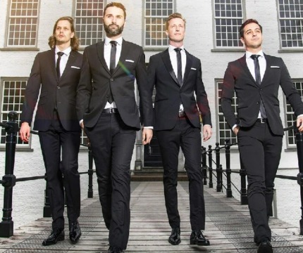 Vier mannen in pakken lopen over een brug voor een wit gebouw met grote ramen, bomen op de achtergrond.