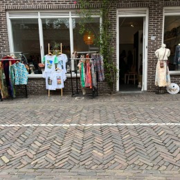 Kledingwinkel met etalages en rekken vol kleurrijke kleding aan de straat, bakstenen gevel en planten.