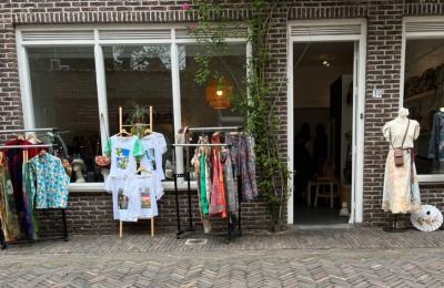 Kledingwinkel met etalages en rekken vol kleurrijke kleding aan de straat, bakstenen gevel en planten.