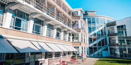 Modern schoolgebouw met grote ramen, een zonnig terras met rode stoelen en tafels onder een blauwe lucht.