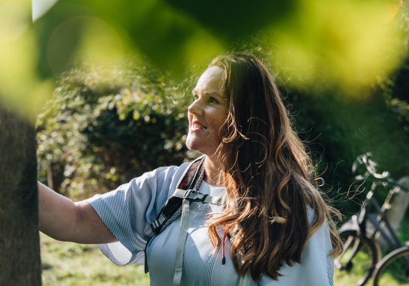 Vrouw glimlacht terwijl ze buiten naast een boom staat, met een fiets op de achtergrond in een zonnige omgeving.