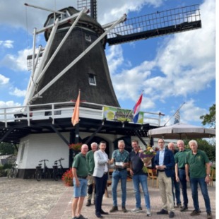 Groepen mensen poseren voor een windmolen en een huis op twee aparte foto’s onder een blauwe lucht.