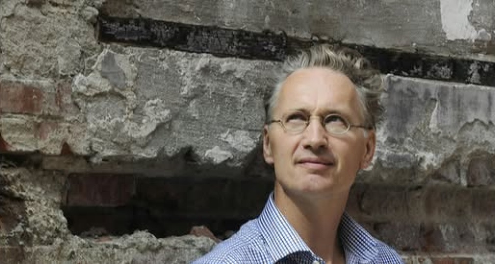 Een man met een bril en grijzend haar poseert voor een oude, verweerde bakstenen muur buiten.