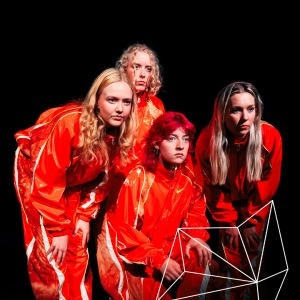 Vier jonge vrouwen in oranje pakken poseren samen op een donker podium voor theaterproductiegroep.