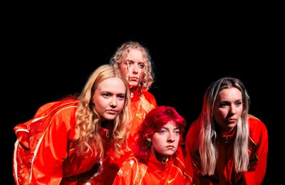 Vier jonge vrouwen in oranje pakken poseren samen op een donker podium voor theaterproductiegroep.