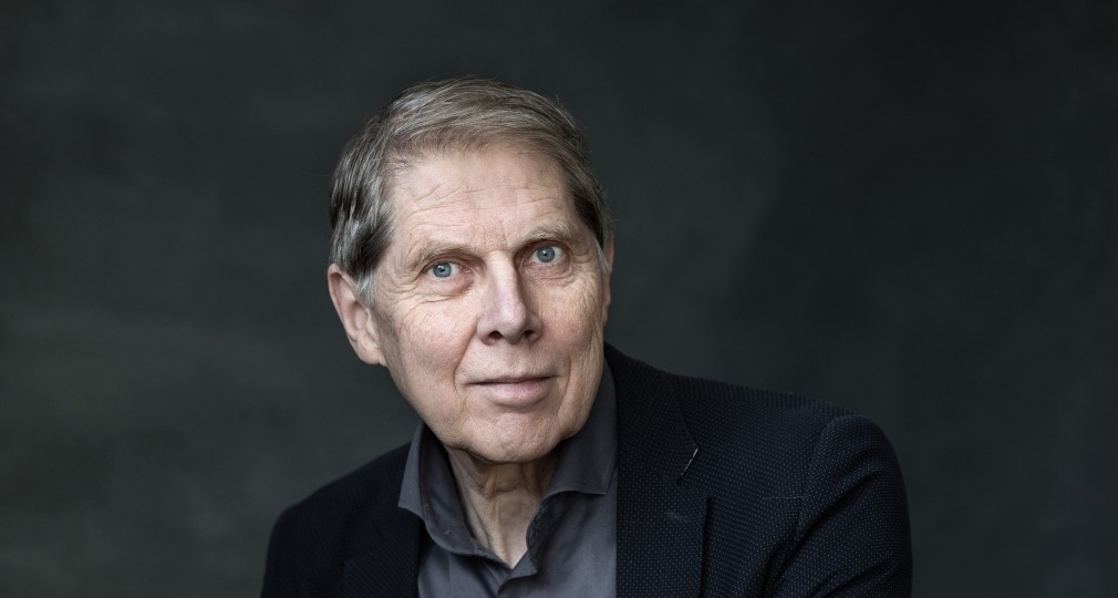 Portret van een oudere man met grijs haar, gekleed in een donker pak en grijs overhemd, handen gevouwen.