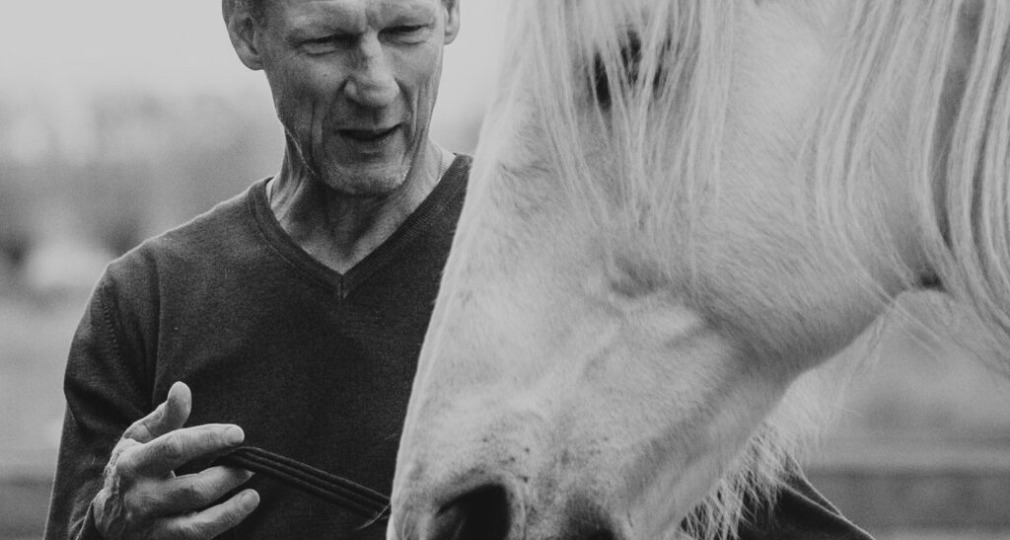 Zwart-witfoto van een man die een wit paard vasthoudt bij zijn teugels, beiden in buitenomgeving.