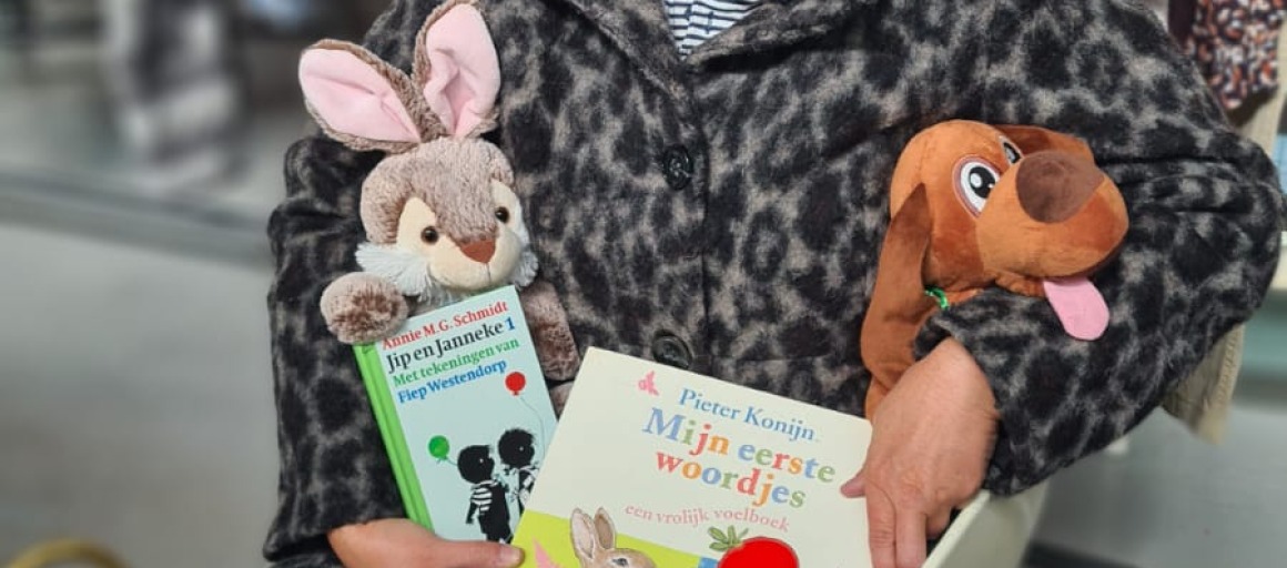 Vrouw met kort haar en dierenknuffels leest kinderboeken in een tweedehandswinkel, omringd door speelgoed.