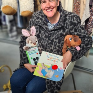 Vrouw met kort haar en dierenknuffels leest kinderboeken in een tweedehandswinkel, omringd door speelgoed.