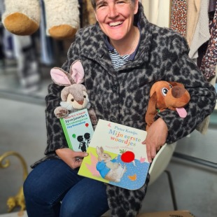 Vrouw met kort haar en dierenknuffels leest kinderboeken in een tweedehandswinkel, omringd door speelgoed.